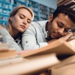Ethnic indian mixed race guy and white girl surrounded by books in library at night. Students are sleeping.