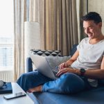 young-attractive-man-sitting-on-sofa-at-home-working-on-laptop-online-using-internet (1)