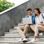 optimistic-couple-man-woman-with-paper-cup-smiling-looking-laptop-while-sitting-stairs-outdoors