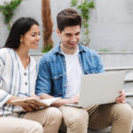 smiling-couple-man-woman-casual-clothes-using-laptop-writing-diary-while-sitting-bench-near-stairs-outdoors