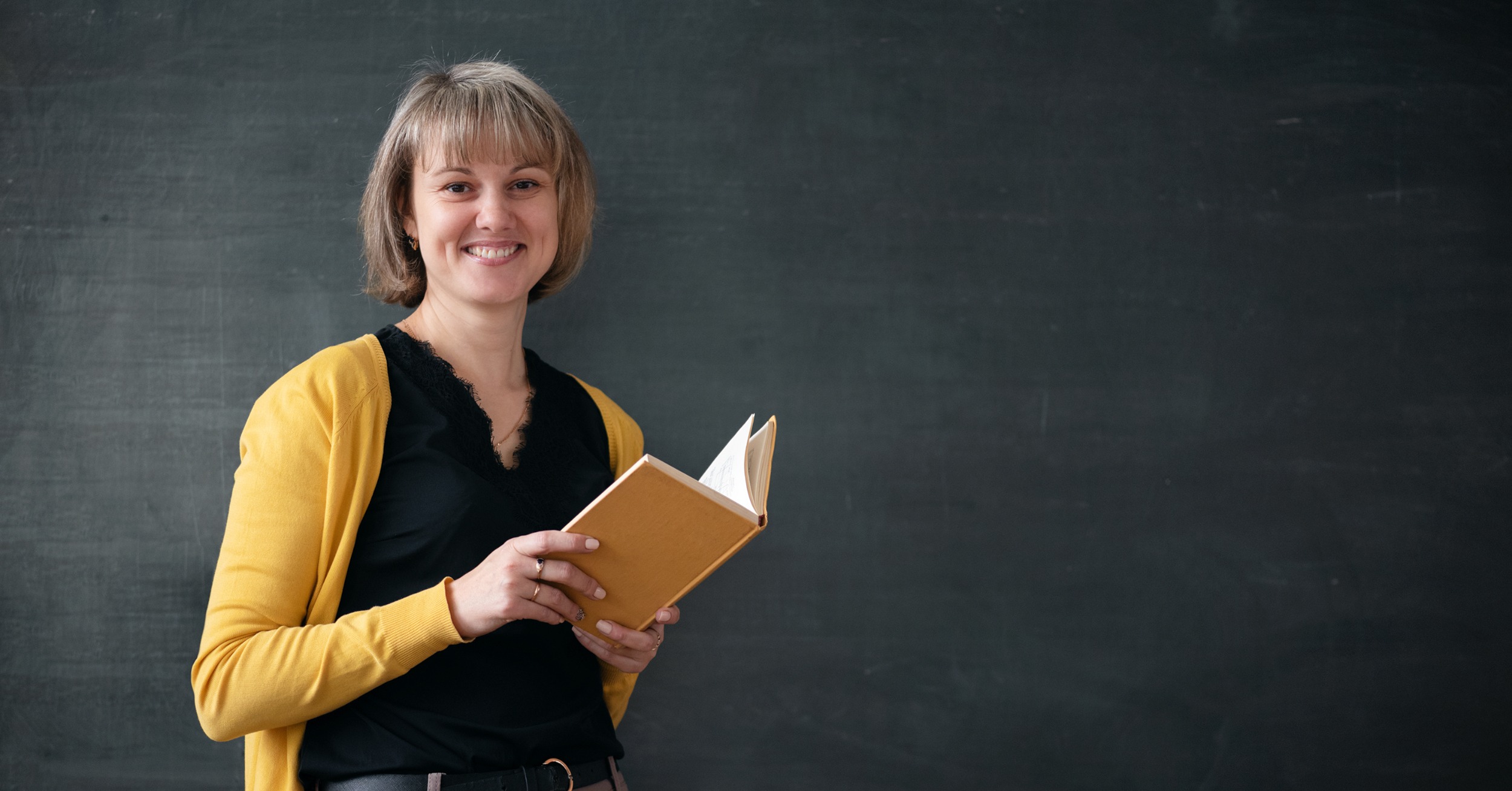 Riconoscimento titolo estero, docente straniera in primo piano con libro in mano e lavagna sullo sfondo